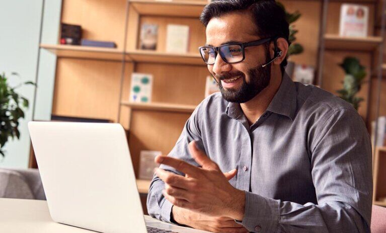 Man wears headset and sits in front of open laptop at desk. A book shelf in background