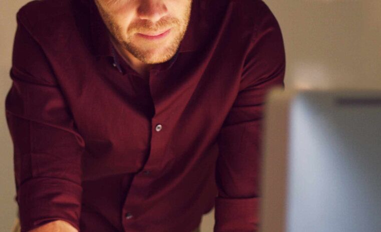 A man standing with his arms on a desk, looking towards a laptop screen | Bachelor of Psychological Science and Criminology