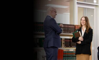 Two Law (PLT) students from ACAP University College standing in front of a bookshelf in an office, talking with and smiling towards a lawyer (2880x1188)