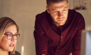 A woman with glasses sitting at, and man standing over a desk looking at a desktop monitor | Bachelor of Psychological Science and Criminology