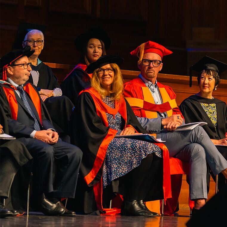 Academic staff sit on stage in traditional and ceremonial robes and caps