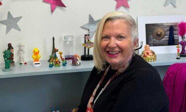 Woman sits in chair in front of desk with trinkets. Stars on wall. Flowers in vase.