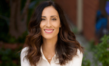 Woman with long brown hair and pink lipstick in a white collared shirt smiles at camera