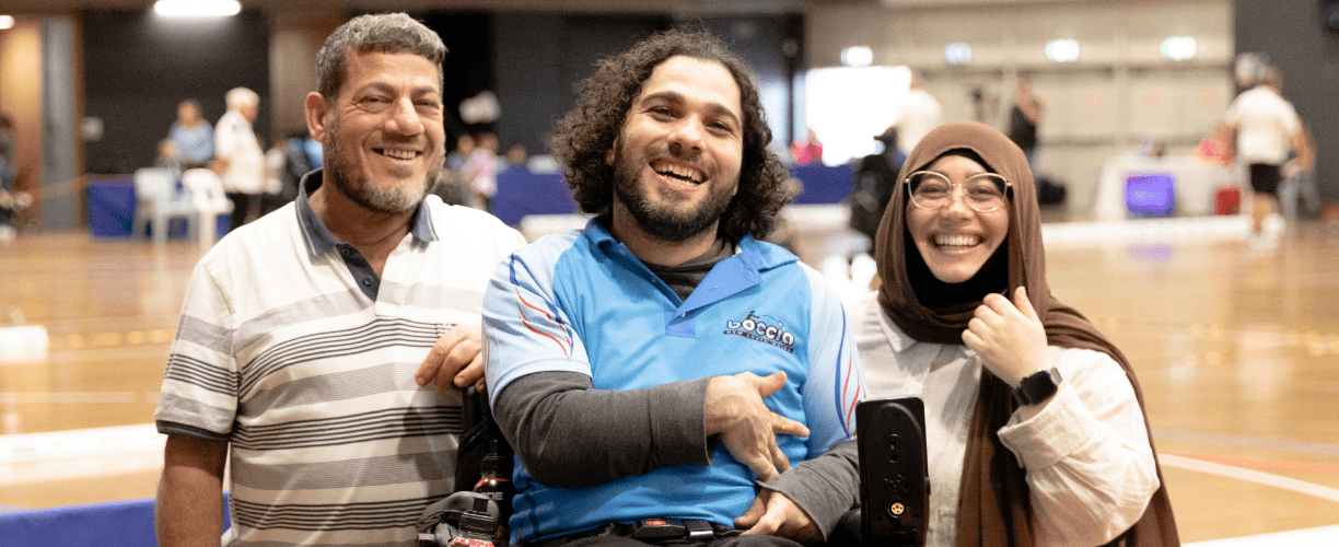two men and a woman wearing a hijab on a sports court smiling. the man in the middle is in a wheelchair