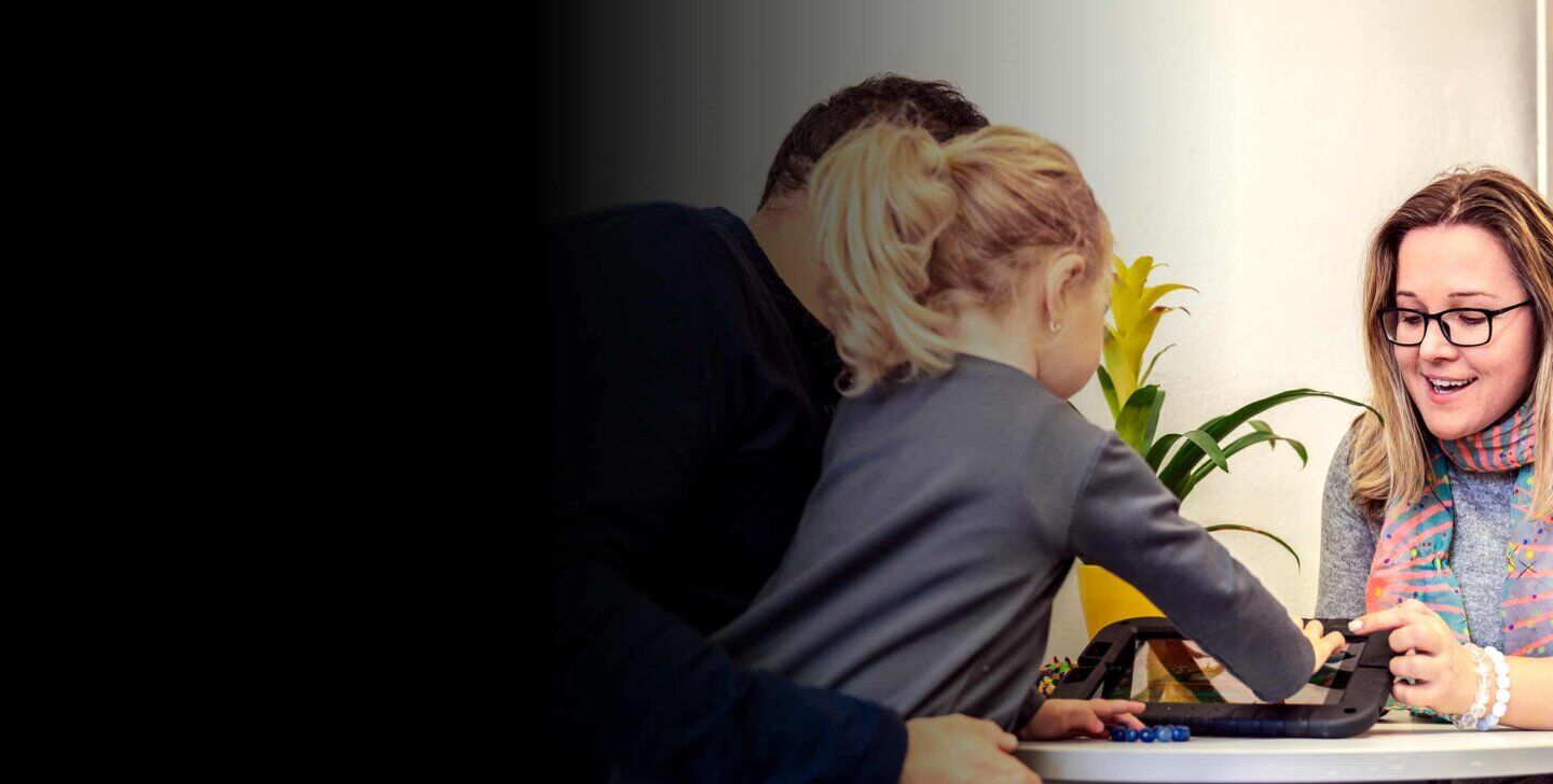 A young child sitting on a man's lap and touching the screen of a tablet, sitting across a table from a female counsellor | Master of Counselling and Psychotherapy