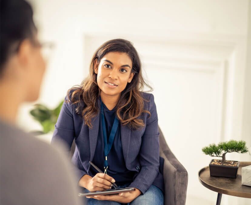 Woman sitting on a chair and facing towards camera with tablet and stylus in hand, looking across at another person in the foreground