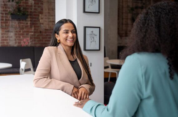 A female management student wearing a light pink suit jacket, sitting at a desk and smiling towards an ACAP lecturer in the foreground (1140x7532)