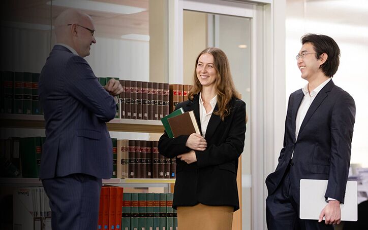 Two Law (PLT) students from ACAP University College standing in front of a bookshelf in an office, talking with and smiling towards a lawyer (724x454)