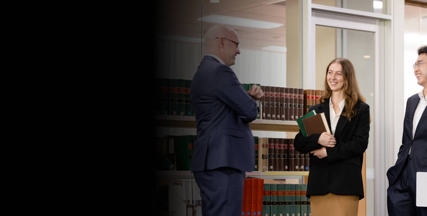 Two Law (PLT) students from ACAP University College standing in front of a bookshelf in an office, talking with and smiling towards a lawyer (2880x1188)
