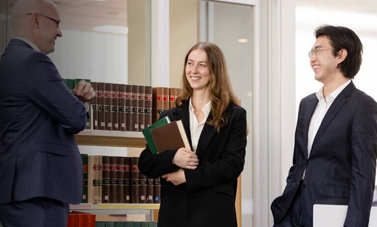 Two Law (PLT) students from ACAP University College standing in front of a bookshelf in an office, talking with and smiling towards a lawyer (828x676)