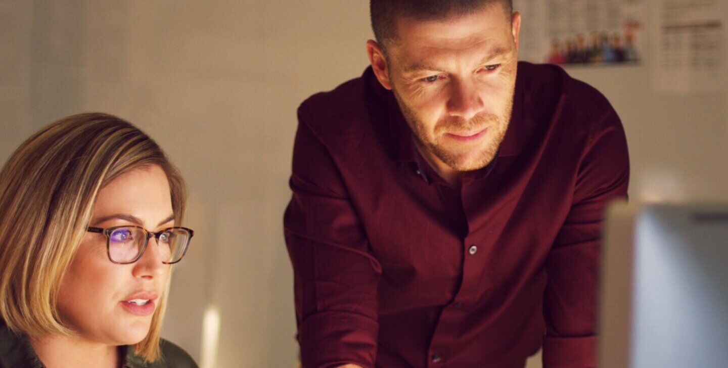 A woman with glasses sitting at, and man standing over a desk looking at a desktop monitor | Bachelor of Psychological Science and Criminology