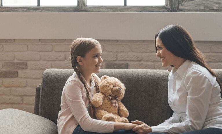 A woman on a couch sits smiles at a young girl also on the couch, holding a teddy bear. She holds her hand as they face each other.