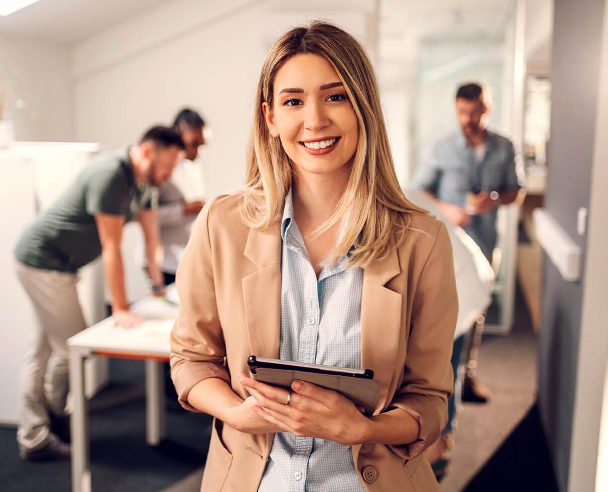 Woman stands in office environment. She is wearing a suit. Holding a tablet. Three men stand around a table in the background