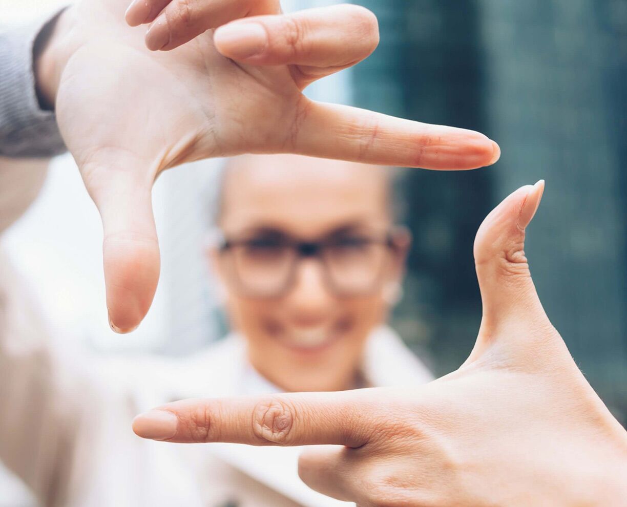 A woman's hands creating a frame with her thumb and index finger, with her face showing through the frame in the background - out of focus.