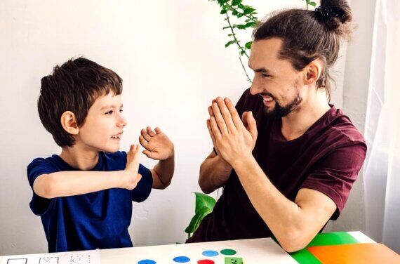 Man with top bun and tee shirt on sits at small table with hands in prayer position. The table has letters and numbers and shapes and coloured paper. He is smiling with young boy who sits opposite him, with his hands up and looking at man.