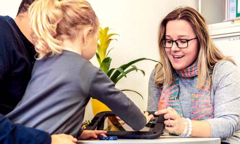 A young child sitting on a man's lap and touching the screen of a tablet, sitting across a table from a female counsellor | Master of Counselling and Psychotherapy