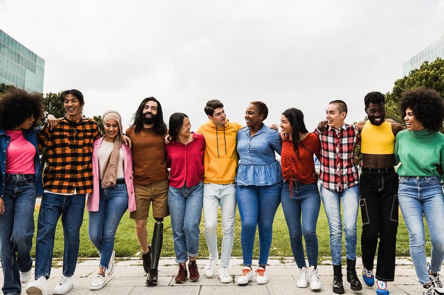 A group of people stand, arm in arm. They all appear young and of mixed ethnicity. They are standing outside, with trees and builds to the left and right of them. They all look at each other, smiling and laughing.