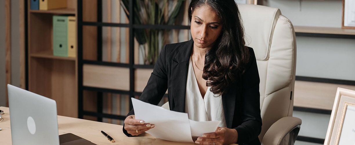Woman sitting at desk in an office chair. She is wearing a suit. A laptop and a closed pen on desk in front of her. She is looking at the papers in her hand. A wall of shelves are behind her.