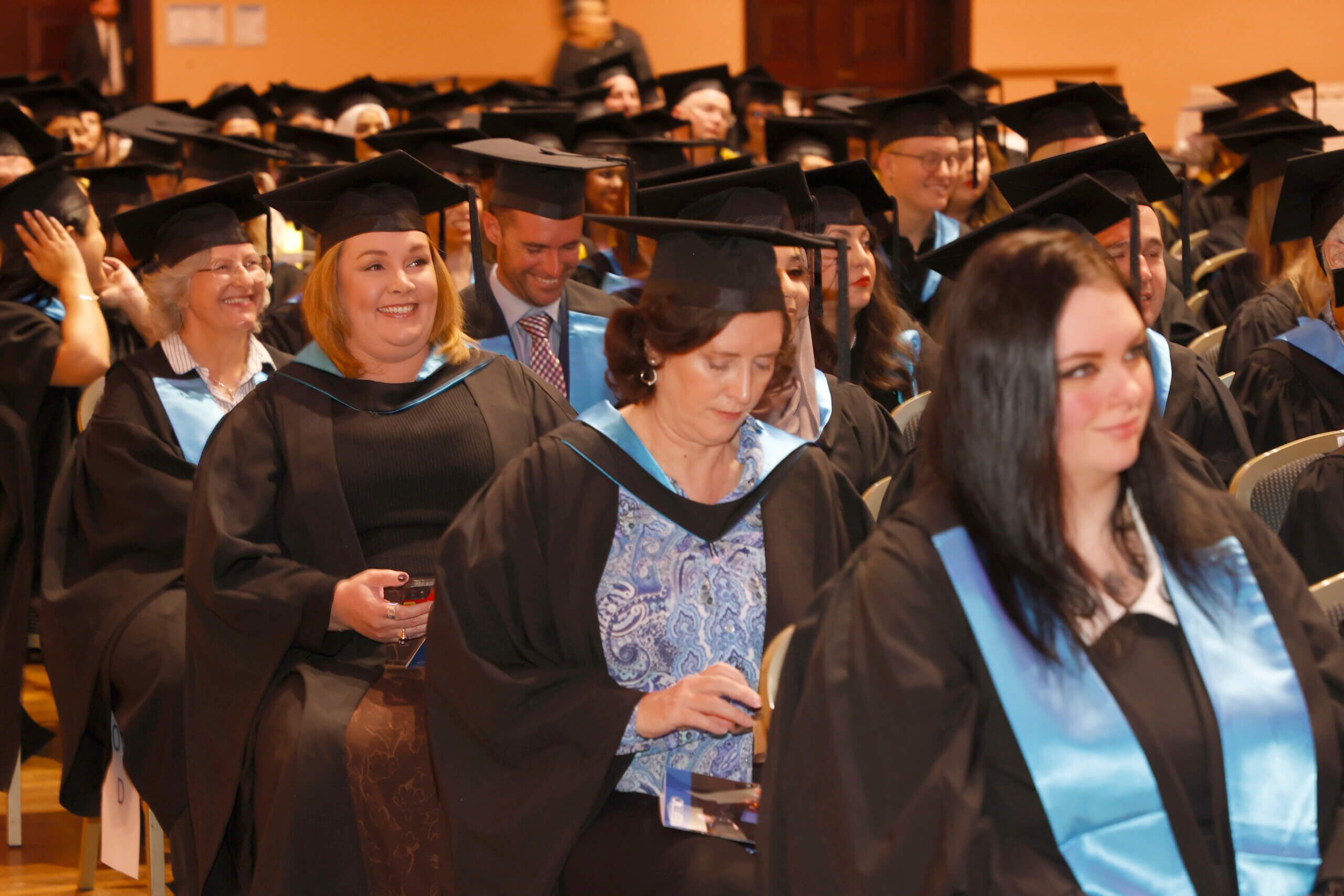 Graduates sitting in hall/large space in ceremonial gowns and caps.