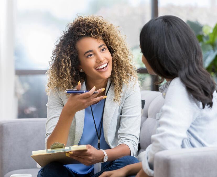 Person speaks to another person on a couch. She holds a pen and a clipboard and has her hand up.