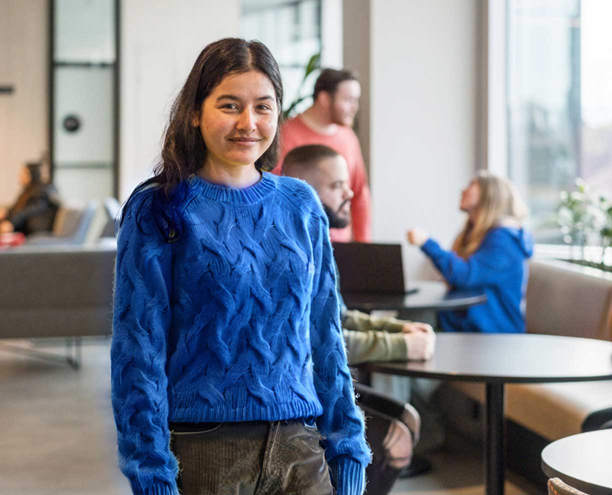 A young woman stands in a room. She wears a blue knit sweater. People sitting in background - talking.
