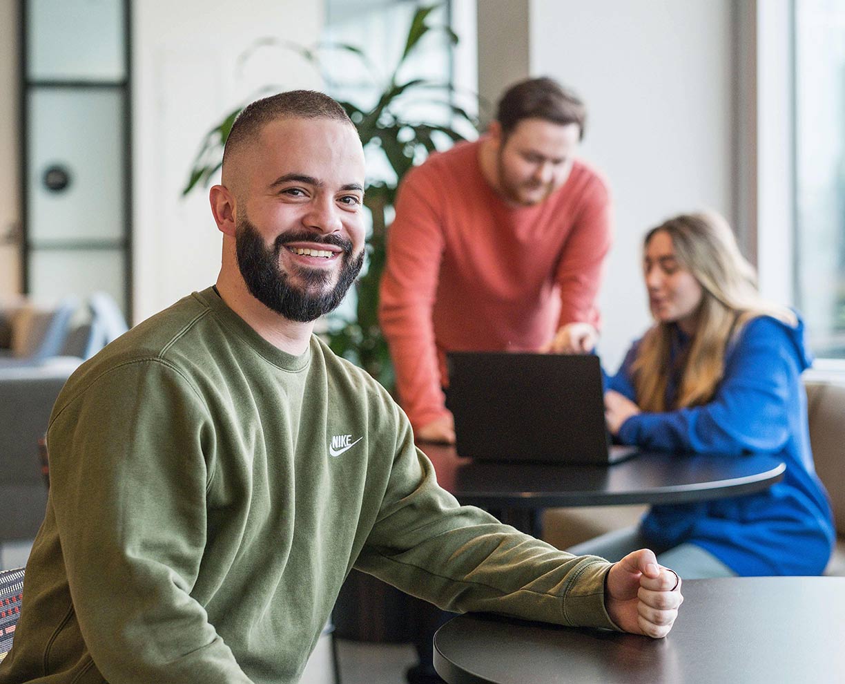 A male student wearing a green sweater, sitting at a table in an ACAP University College common area in front of two other students in the background (1224x990)