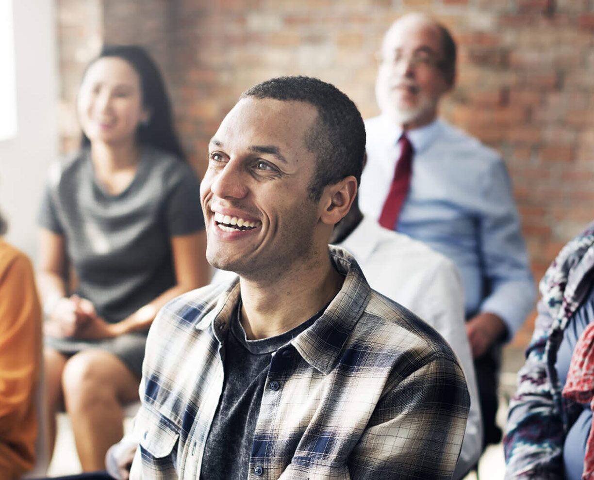 A smiling man sitting at an ACAP Clinic event, with other people sitting in the background in front of an exposed brick wall (1224x990)